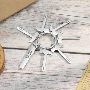 May include: A silver keychain with a saw blade charm that reads "Best Dad I Ever Saw". The keychain has multiple hammer charms with names engraved on each one: Nathan, Kelya, Cesar, Eileen, Maylynn, and Zoriah.