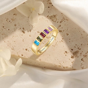 Puede incluir: Un anillo dorado con seis gemas rectangulares de colores. El anillo está grabado con el texto "Happy Mother's Day!". Las gemas son azules, verdes, rojas, moradas y rosas. El anillo se muestra sobre una superficie texturizada.
