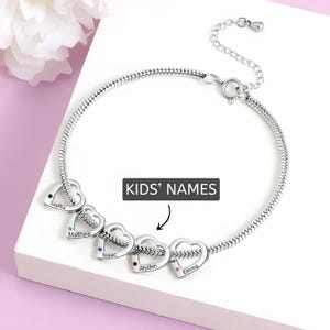 May include: A silver bracelet with heart-shaped charms, each engraved with a name and a coloured gemstone. The bracelet has a snake chain design and is labelled "KIDS' NAMES".