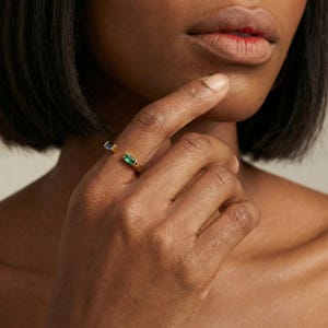 May include: A gold-colored open ring featuring a rectangular emerald-green gemstone and a rectangular blue gemstone. The ring is worn on a finger, with the gemstones positioned on either side of the opening.