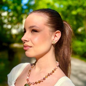 Puede incluir: Un collar con colgante de corazón dorado y una cadena de cuentas de piedras rosas y amarillas. Se ven pendientes dorados a juego. La joya se lleva con un top color crema con estampado floral.