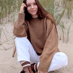 Può includere: Una persona indossa un maglione marrone, pantaloni bianchi e stivali marroni. Il maglione ha un polsino a coste e una vestibilità ampia. Gli stivali hanno una cerniera laterale e lacci neri. La persona è seduta sulla sabbia.