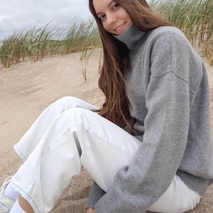 Può includere: Una persona indossa un dolcevita grigio e pantaloni bianchi seduta su una spiaggia sabbiosa. Il maglione ha colletto e polsini a coste. I pantaloni sono di colore chiaro. Lo sfondo include erba di spiaggia e un cielo nuvoloso.