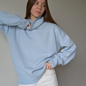 May include: A light blue turtleneck sweater with long sleeves. The sweater is paired with white jeans. The model is wearing the sweater and jeans in front of a gray background.