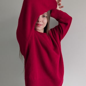May include: A long-sleeved, deep red knit sweater featuring a classic crew neckline and ribbed cuffs at the wrists and hem. The garment is constructed with a solid-colored, fine-gauge knit texture, providing a consistent appearance throughout the piece.