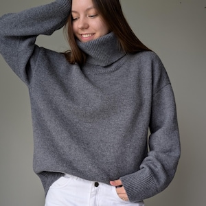 May include: A gray turtleneck sweater paired with white jeans. The sweater has a ribbed collar and cuffs. The model is smiling and has their arm raised. The jeans have a button closure and pockets.