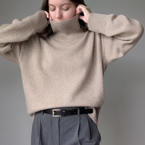Puede incluir: Un su&eacute;ter beige de cuello alto con mangas largas y un cintur&oacute;n de cuero negro. El su&eacute;ter est&aacute; hecho de una tela suave y de punto y tiene un ajuste holgado.