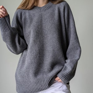 Może przedstawiać: Szary, oversize'owy sweter z okrągłym dekoltem i długimi rękawami. Sweter ma żebrowane wykończenie u dołu i przy mankietach.