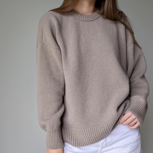 May include: A tan-colored, crew neck sweater with ribbed cuffs and hem. The sweater has a relaxed fit and long sleeves. The material appears to be a medium-weight knit, suitable for cooler weather. The sweater is paired with white pants.