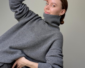 Puede incluir: Un suéter de punto gris carbón con cuello alto, mangas largas con puños acanalados y un corte holgado y extragrande. La prenda está confeccionada con un material de mezcla de lana suave y texturizado con una apariencia jaspeada uniforme.