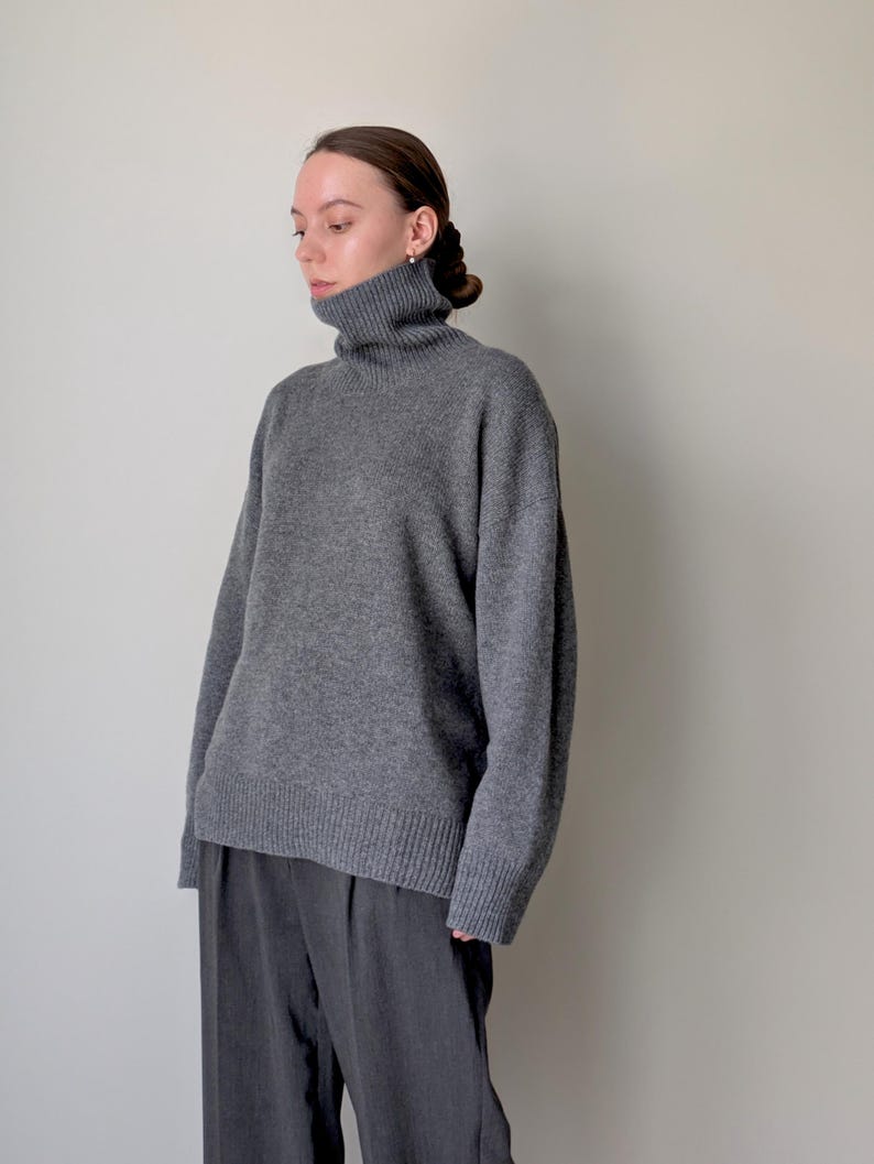Op de afbeelding: Een antracietgrijze trui met lange mouwen, een hoge col, verlaagde schouders en geribbelde gebreide details bij de manchetten en de zoom, gecombineerd met een donkergrijze broek met een zichtbare verticale vouw aan de voorzijde.