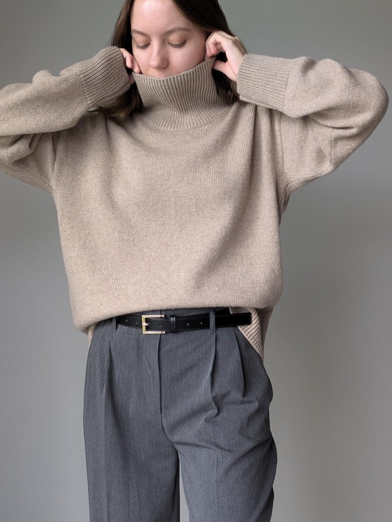 Puede incluir: Un su&eacute;ter beige de cuello alto con mangas largas y un cintur&oacute;n de cuero negro. El su&eacute;ter est&aacute; hecho de una tela suave y de punto y tiene un ajuste holgado.
