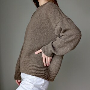 May include: A long-sleeved, brown knit sweater featuring a crew neckline, dropped shoulders, and ribbed cuffs and hem. The sweater has a relaxed fit and a textured, solid-colored knit pattern, paired with plain white trousers.