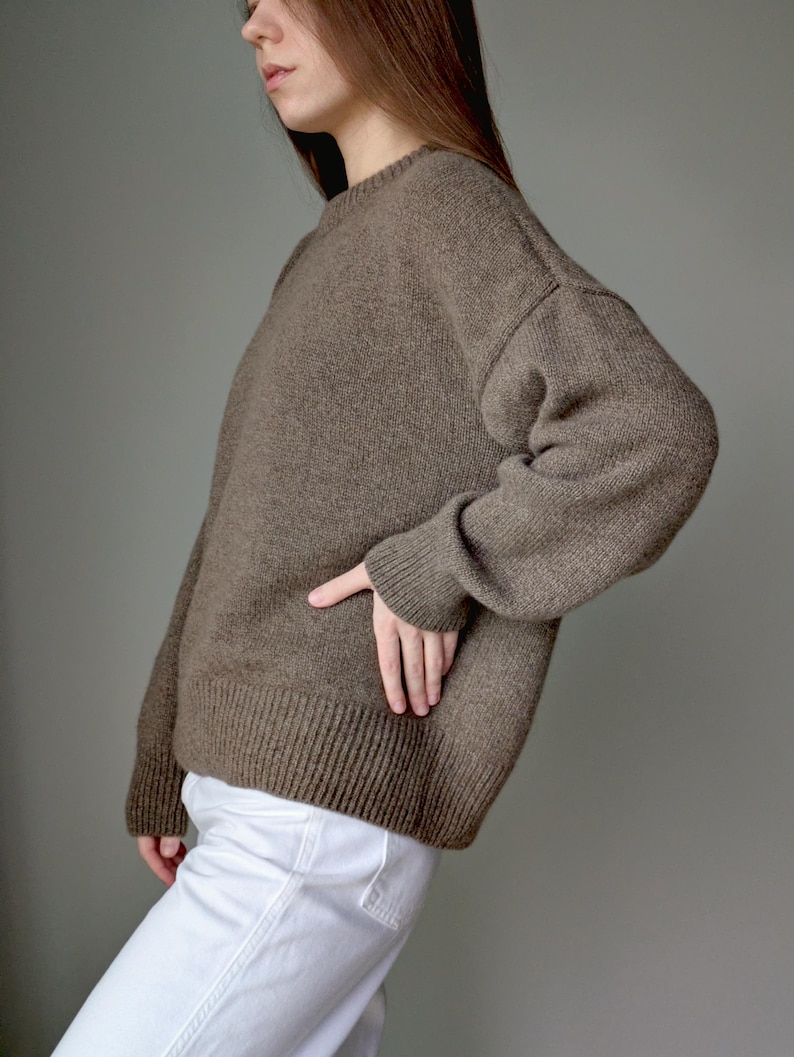 May include: A long-sleeved, brown knit sweater featuring a crew neckline, dropped shoulders, and ribbed cuffs and hem. The sweater has a relaxed fit and a textured, solid-colored knit pattern, paired with plain white trousers.