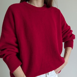 May include: A vibrant red long-sleeve sweater featuring a classic crew neckline and ribbed knit cuffs at the wrists and hem. The garment has a relaxed, loose-fitting silhouette and is paired with white denim pants with a visible metal button.