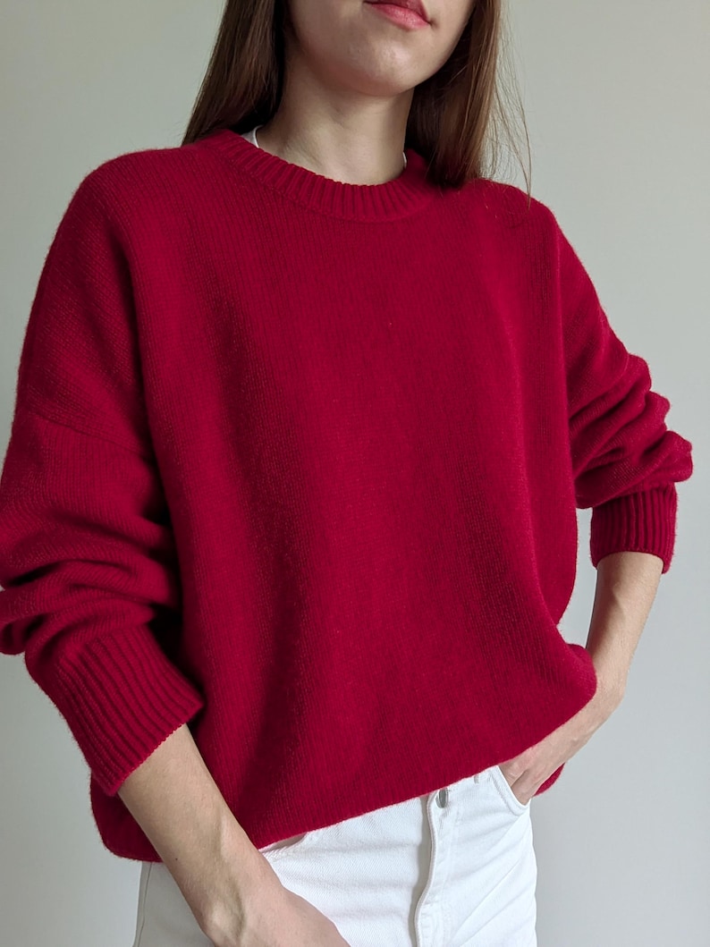 May include: A vibrant red long-sleeve sweater featuring a classic crew neckline and ribbed knit cuffs at the wrists and hem. The garment has a relaxed, loose-fitting silhouette and is paired with white denim pants with a visible metal button.