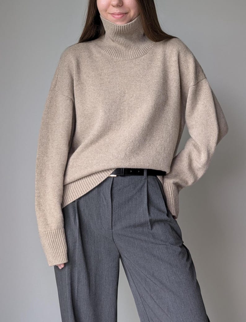 Puede incluir: Un su&eacute;ter beige de cuello alto con mangas largas y un corte holgado. El su&eacute;ter se combina con pantalones grises y un cintur&oacute;n negro.