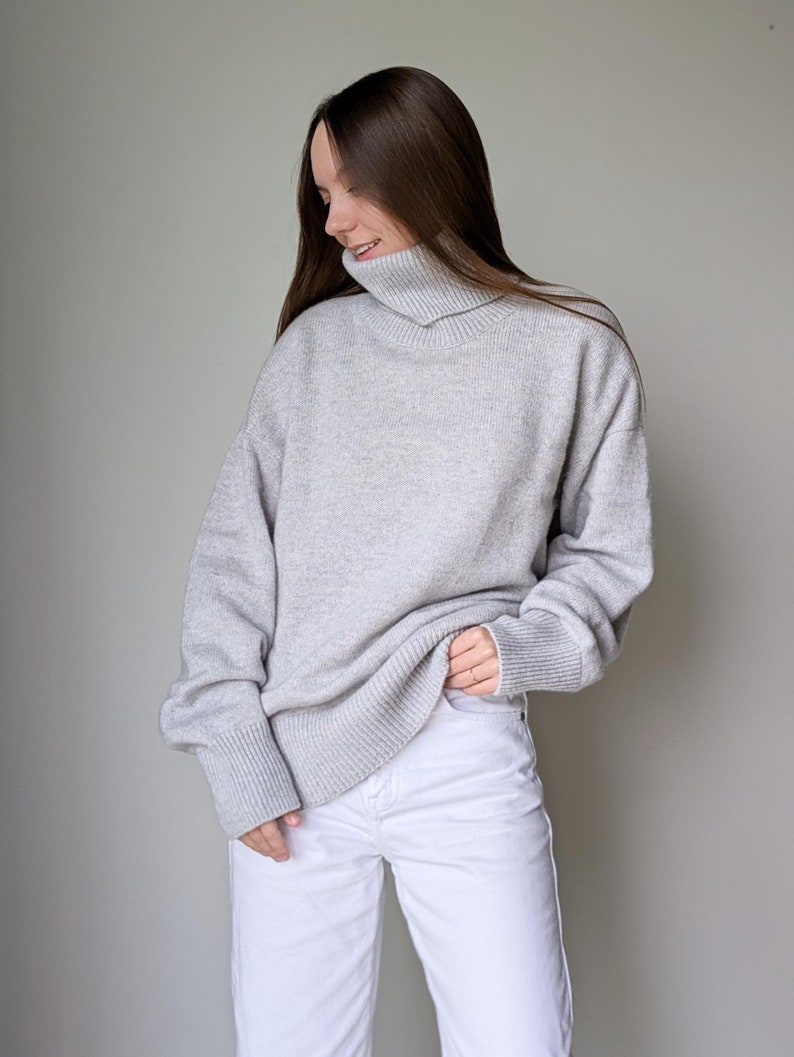 May include: A light gray turtleneck sweater with ribbed cuffs and hem. The sweater is paired with white jeans. The model is smiling and looking down. The background is a solid light gray.