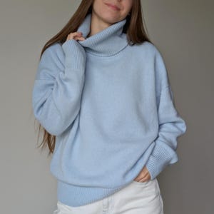 May include: A light blue turtleneck sweater with long sleeves. The sweater has a ribbed collar and cuffs. The person is wearing white jeans. The sweater is a soft, comfortable material.