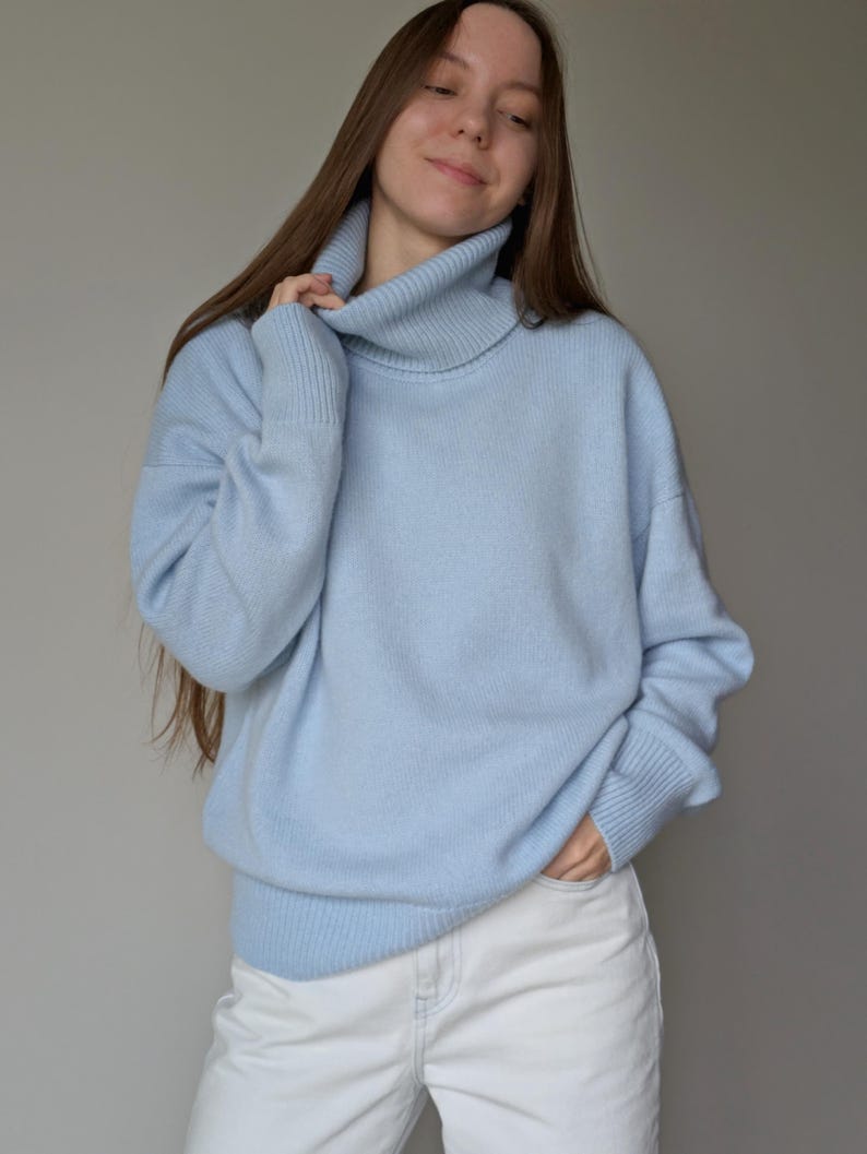 May include: A light blue turtleneck sweater with long sleeves. The sweater has a ribbed collar and cuffs. The person is wearing white jeans. The sweater is a soft, comfortable material.