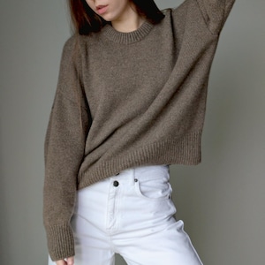 May include: A long-sleeved, brown knit sweater featuring a ribbed crew neckline, ribbed cuffs, and a ribbed hem. The sweater has a relaxed fit with dropped shoulders and is paired with white denim pants that feature a button fly and front pockets.