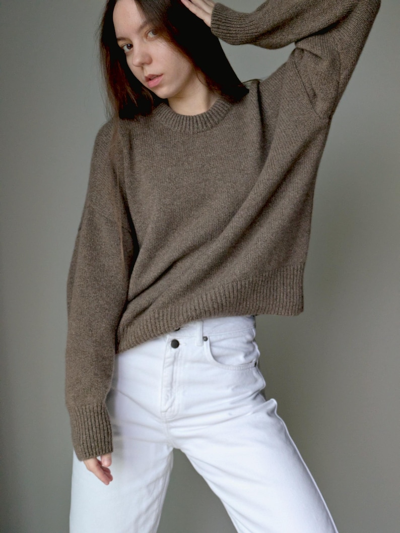 May include: A long-sleeved, brown knit sweater featuring a ribbed crew neckline, ribbed cuffs, and a ribbed hem. The sweater has a relaxed fit with dropped shoulders and is paired with white denim pants that feature a button fly and front pockets.