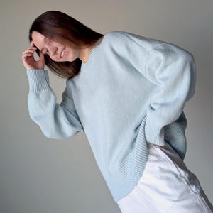 May include: A light blue long-sleeved sweater with a ribbed crew neckline, ribbed cuffs, and a ribbed hem. The sweater features a relaxed fit with dropped shoulders and a solid, plain knit texture throughout the garment, paired with white trousers.