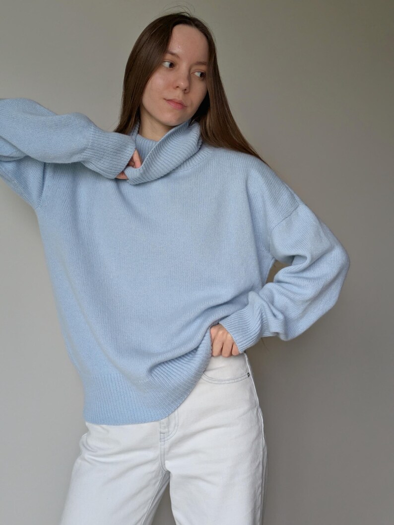 May include: A light blue turtleneck sweater with long sleeves. The sweater is paired with white jeans. The model is wearing the sweater and jeans in front of a gray background.