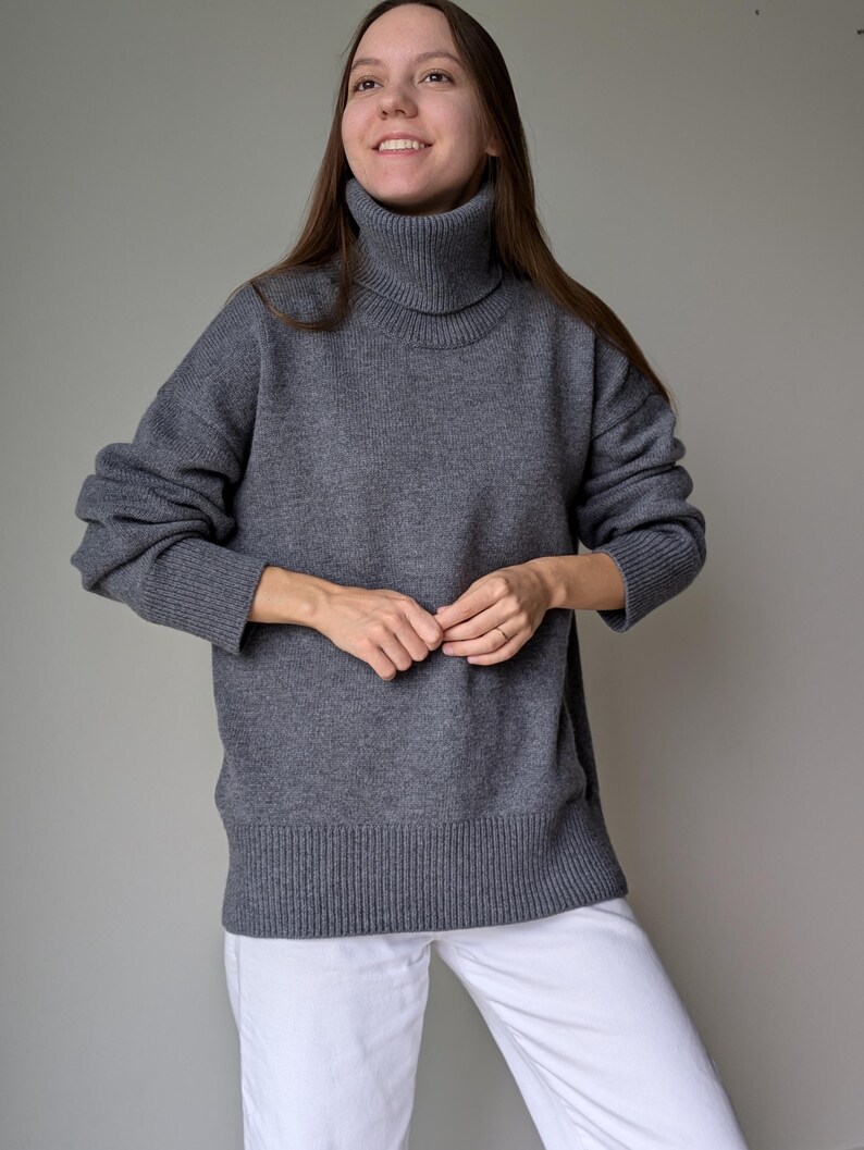 May include: A gray turtleneck sweater with long sleeves. The sweater has a ribbed hem and cuffs. The person is wearing white pants. The sweater is a solid color and appears to be made of a soft knit material.