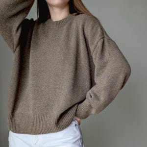 May include: A long-sleeved, taupe-colored knit sweater featuring a classic crew neckline and ribbed cuffs at the wrists and hem. The garment is paired with plain white denim pants, creating a neutral-toned ensemble with a simple, clean aesthetic.