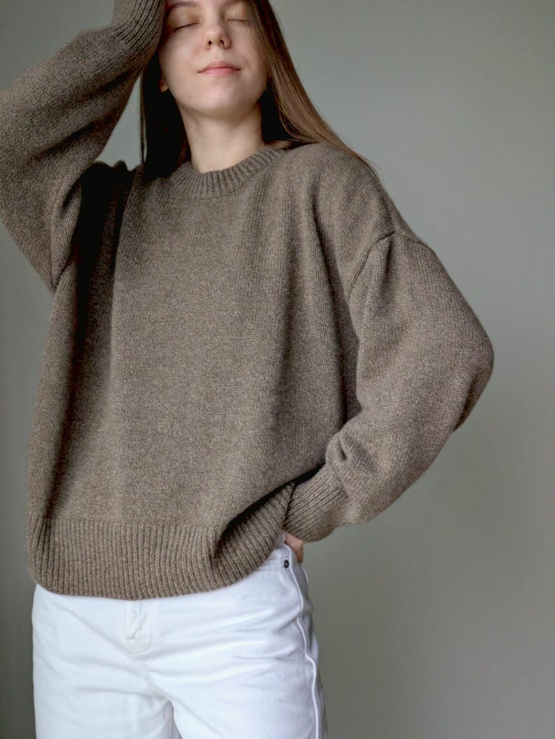 May include: A long-sleeved, taupe-colored knit sweater featuring a classic crew neckline and ribbed cuffs at the wrists and hem. The garment is paired with plain white denim pants, creating a neutral-toned ensemble with a simple, clean aesthetic.