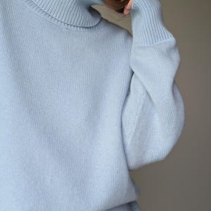 May include: A light blue turtleneck sweater. The sweater has a ribbed collar and cuffs, and a soft, knitted texture. The sleeves are long and the overall style is casual and comfortable. The color is a pale, pastel blue.