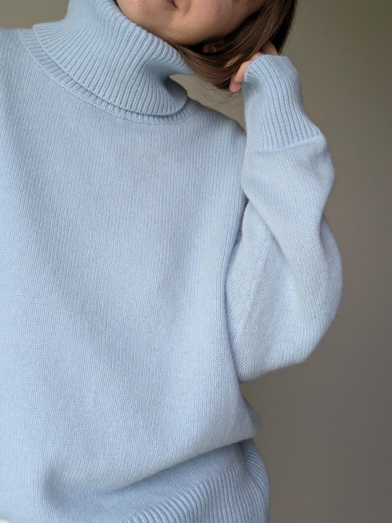 May include: A light blue turtleneck sweater. The sweater has a ribbed collar and cuffs, and a soft, knitted texture. The sleeves are long and the overall style is casual and comfortable. The color is a pale, pastel blue.