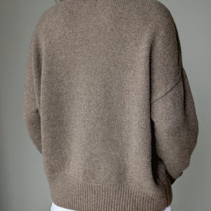 May include: A long-sleeved, crew-neck sweater in a solid taupe-brown color with a ribbed knit texture at the collar, cuffs, and hemline. The garment features dropped shoulders and a relaxed fit, paired with plain white trousers.