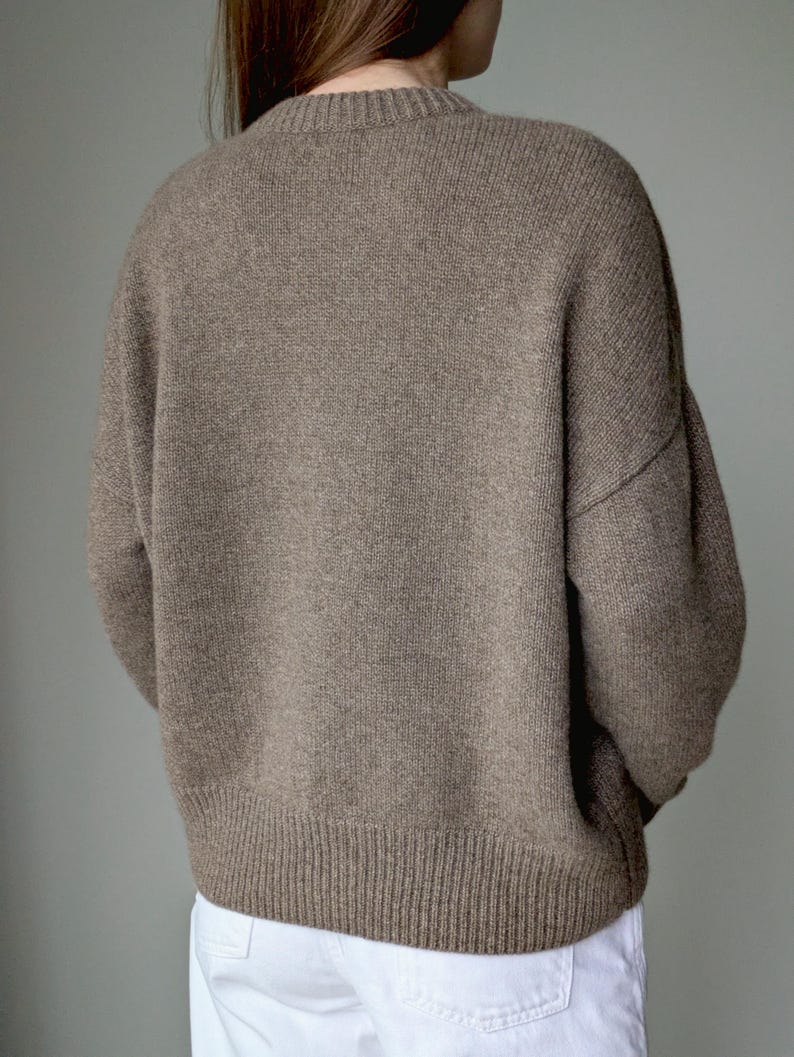 May include: A long-sleeved, crew-neck sweater in a solid taupe-brown color with a ribbed knit texture at the collar, cuffs, and hemline. The garment features dropped shoulders and a relaxed fit, paired with plain white trousers.