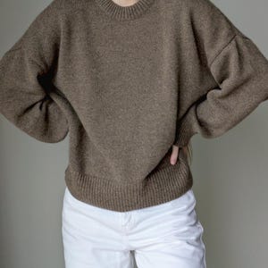 May include: A brown long-sleeve sweater with a crew neckline and ribbed cuffs and hem, paired with white pants. The sweater features a relaxed fit with dropped shoulders and a textured knit construction, providing a solid neutral tone throughout.