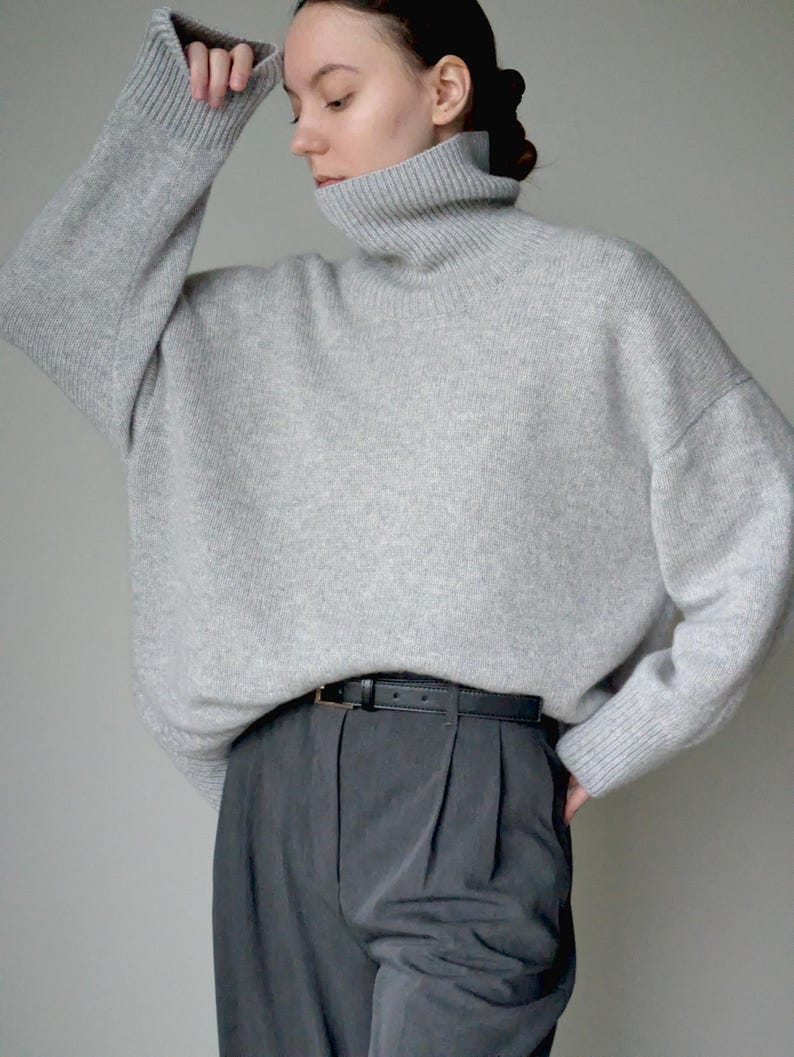Op de afbeelding: Een lichtgrijze oversized coltrui met lange mouwen en geribbelde boorden, gecombineerd met een donkergrijze pantalon met plooien en een smalle zwarte leren riem met een kleine goudkleurige gesp, wat zorgt voor een neutraal ensemble.