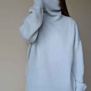May include: A light blue turtleneck sweater. The sweater has a ribbed hem and cuffs, and a wide, oversized turtleneck collar. The model is wearing the sweater and white pants. The sweater is made of a soft, knitted material.