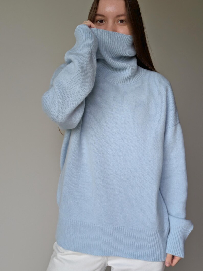 May include: A light blue turtleneck sweater. The sweater has a ribbed hem and cuffs, and a wide, oversized turtleneck collar. The model is wearing the sweater and white pants. The sweater is made of a soft, knitted material.