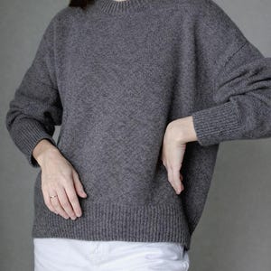 May include: A charcoal gray long-sleeve sweater with a ribbed crew neckline, ribbed cuffs, and a ribbed hem. The sweater features a relaxed fit with dropped shoulders and a textured knit construction, paired with plain white trousers.