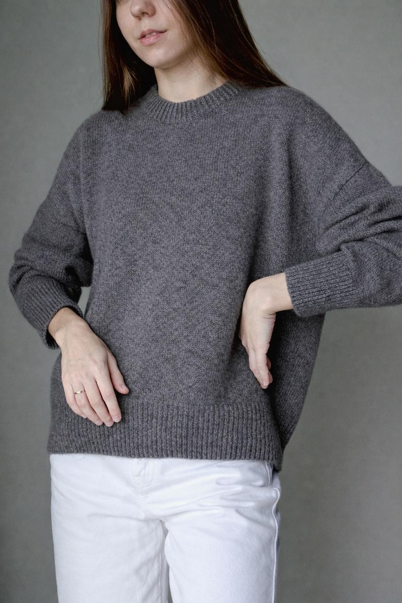 May include: A charcoal gray long-sleeve sweater with a ribbed crew neckline, ribbed cuffs, and a ribbed hem. The sweater features a relaxed fit with dropped shoulders and a textured knit construction, paired with plain white trousers.