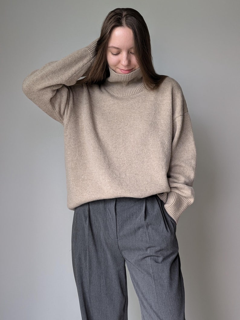 Puede incluir: Un su&eacute;ter beige de cuello alto con mangas largas y un corte holgado. El su&eacute;ter se combina con pantalones grises plisados.