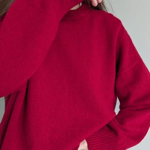 May include: A long-sleeved, solid deep red sweater featuring a ribbed crew neckline, ribbed cuffs, and a ribbed hem. The knit garment has a relaxed fit with dropped shoulders and a consistent, fine-textured weave throughout the entire piece.