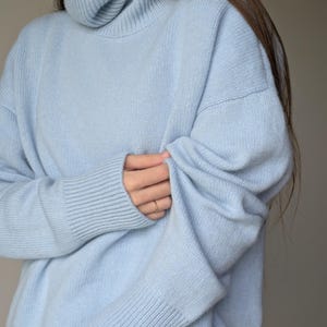 May include: A light blue turtleneck sweater. The sweater has a ribbed collar and cuffs, and a relaxed fit. The sleeves are long, and the material appears soft. The overall style is casual and comfortable, suitable for cooler weather.