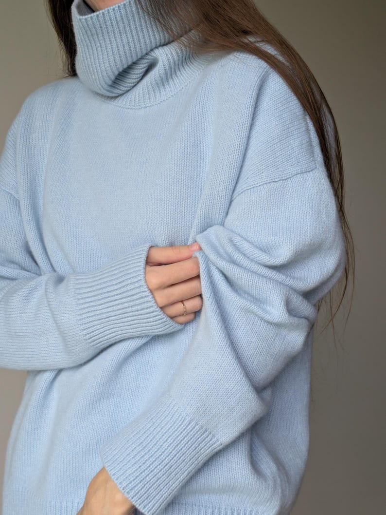 May include: A light blue turtleneck sweater. The sweater has a ribbed collar and cuffs, and a relaxed fit. The sleeves are long, and the material appears soft. The overall style is casual and comfortable, suitable for cooler weather.
