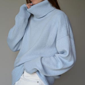 May include: A light blue turtleneck sweater. The sweater has long sleeves and a ribbed collar. The material appears soft and comfortable. The sweater is paired with white pants. The overall style is casual and relaxed.