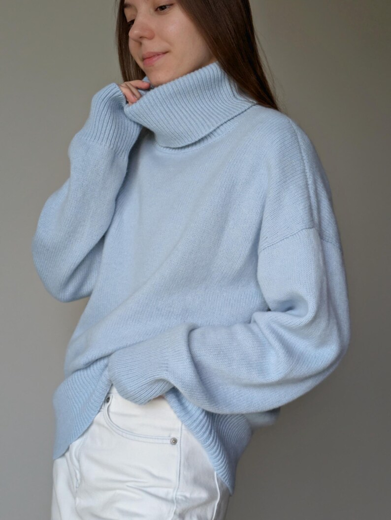 May include: A light blue turtleneck sweater. The sweater has long sleeves and a ribbed collar. The material appears soft and comfortable. The sweater is paired with white pants. The overall style is casual and relaxed.