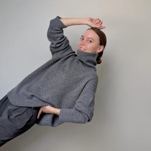 Op de afbeelding: Een antracietgrijze gebreide trui met een hoge col, lange mouwen met geribbelde boorden en een relaxte pasvorm. Het kledingstuk wordt gecombineerd met een donkergrijze broek met een zichtbaar plooidetail bij de tailleband.