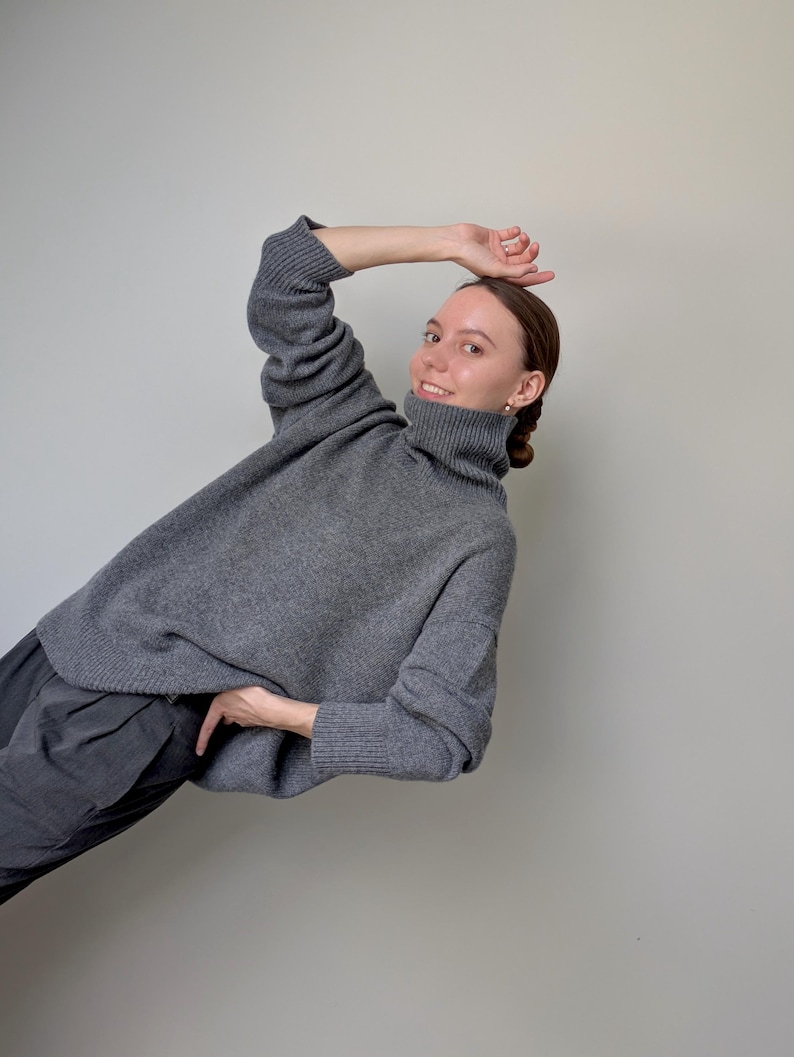 Op de afbeelding: Een antracietgrijze gebreide trui met een hoge col, lange mouwen met geribbelde boorden en een relaxte pasvorm. Het kledingstuk wordt gecombineerd met een donkergrijze broek met een zichtbaar plooidetail bij de tailleband.