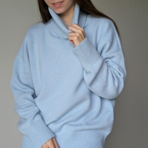 May include: A light blue sweater with a turtleneck collar and ribbed cuffs. The sweater has a relaxed fit and is made of a soft material. The model is wearing white pants. This is a casual, everyday sweater.